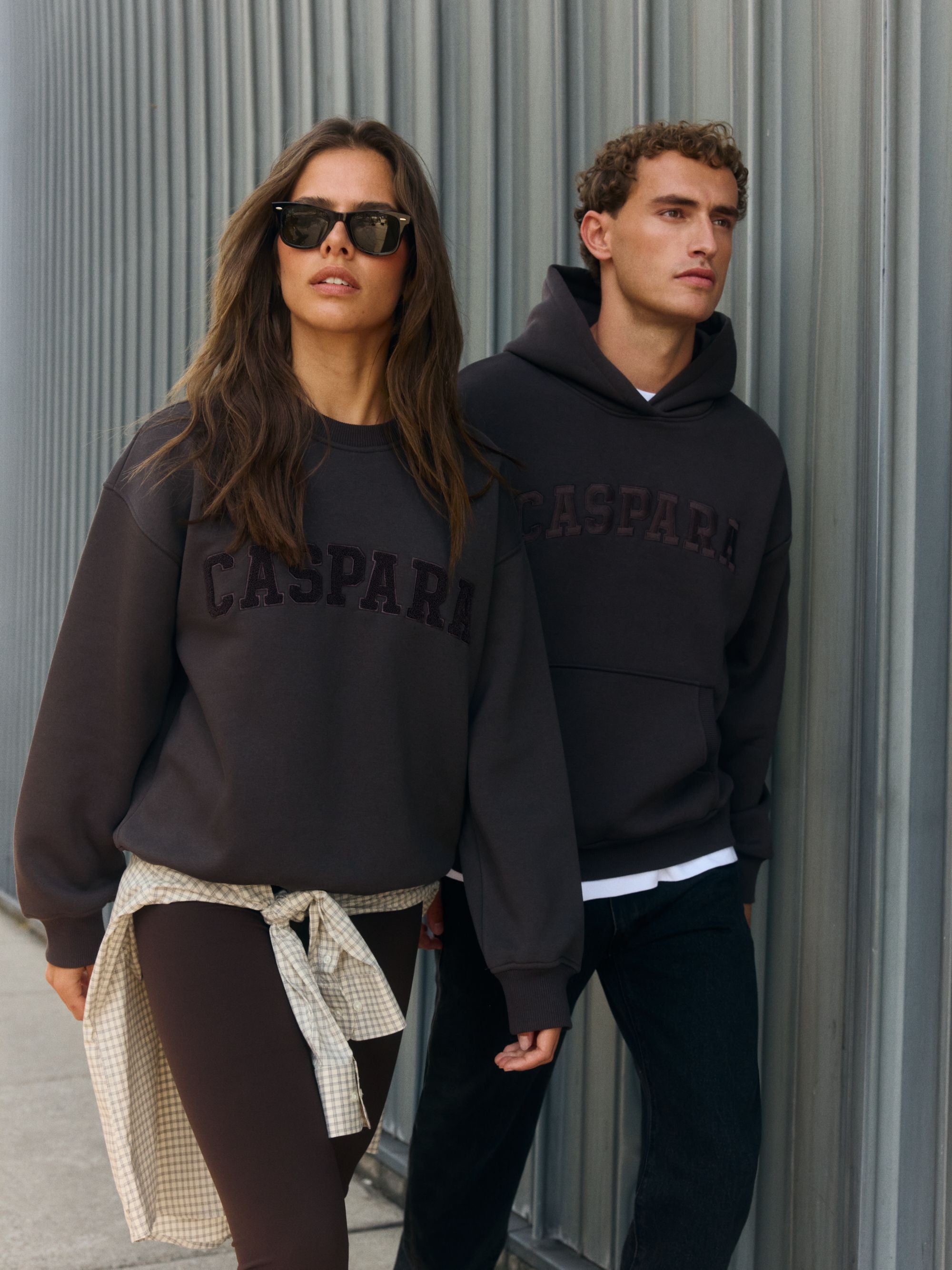Man wearing an espresso CASPARA logo hoodie and woman in a matching sweatshirt outdoors.